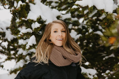 A woman in winter clothes smiles amongst evergreen trees covered in snow.