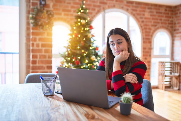 Beautiful woman sitting at the table working with laptop at home around christmas tree thinking look...