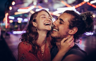 A happy couple laughs in an amusement park on a date at night with lights faded in the background.