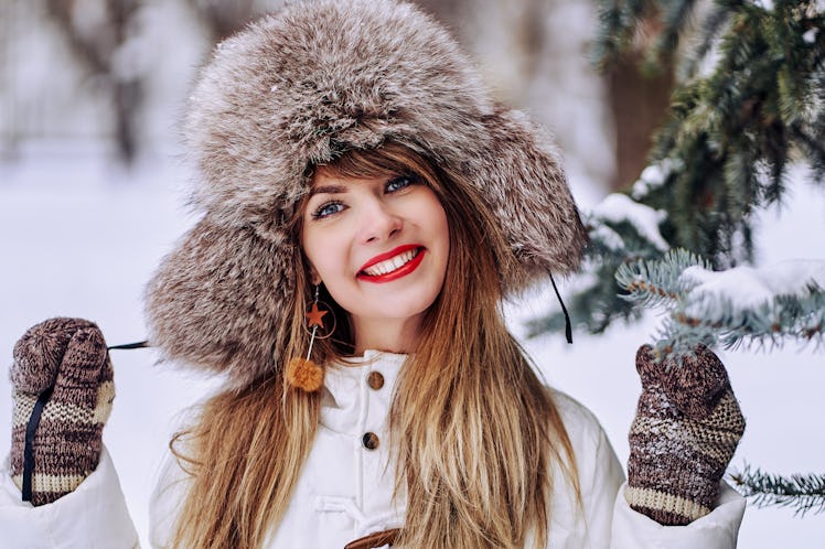 A brunette woman wearing mittens and a matching hat smiles next to an evergreen tree in winter.