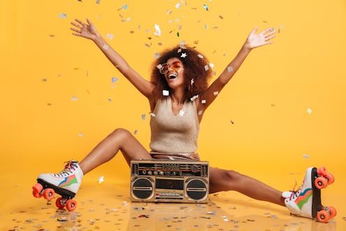 Charming young african woman in retro wear and roller scates throwing confetti while sitting with bo...