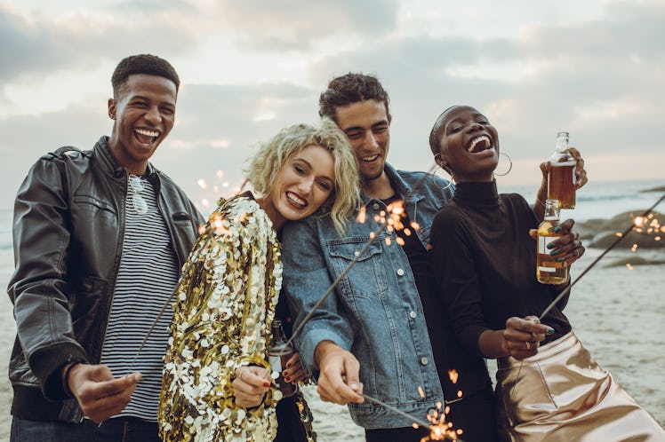 A group of friends in sparkly and stylish clothes holds sparklers and bottles of beer while celebrat...