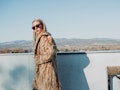 A blonde woman in a long coat and sunglasses stands on a balcony overlooking an underrated wintry to...