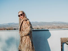 A blonde woman in a long coat and sunglasses stands on a balcony overlooking an underrated wintry to...