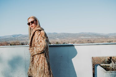 A blonde woman in a long coat and sunglasses stands on a balcony overlooking an underrated wintry to...