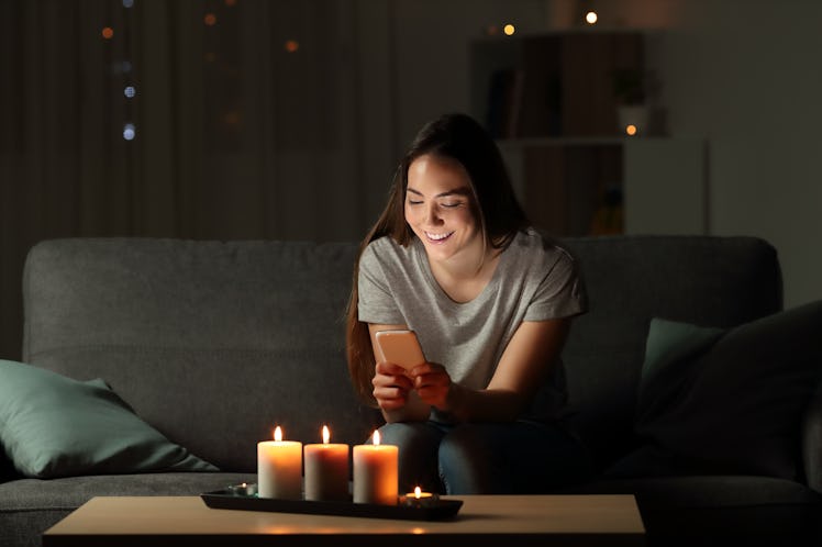 Woman using a smart phone in the night with candle lights sitting on a couch in the living room at h...