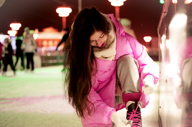 A woman in a bright pink jacket ties her ice skates while skating at a rooftop ice skating rink.