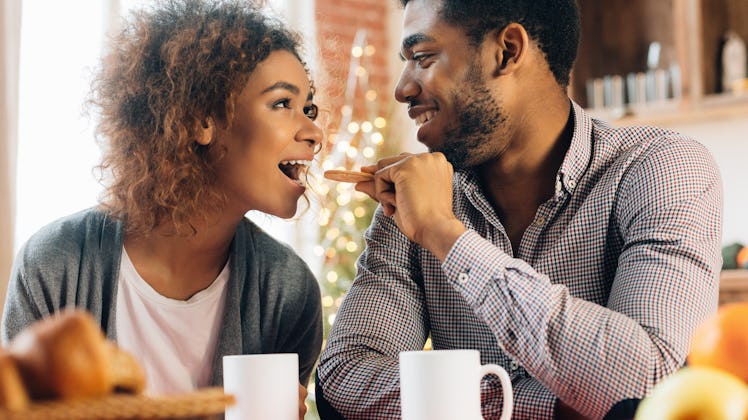 A happy couple sits at a kitchen table with coffee mugs, while one feeds the other a cookie.