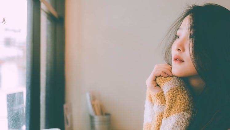 Beautiful young girl Black hair, yellow sweater sitting alone sad at home look out window sad Winter...