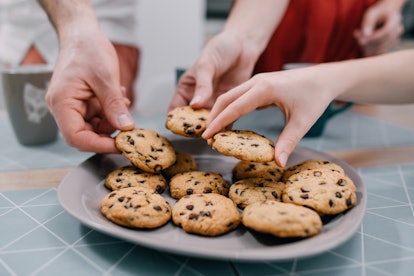 Three hands take fresh chocolate chip cookies from plate.