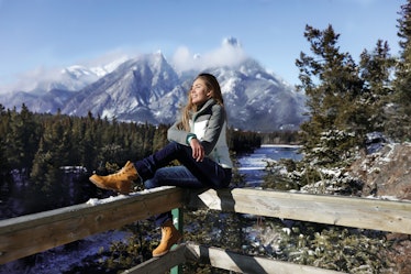 Sports girl sitting on a wooden fence, wearing sportswear, jeans and boots. Cold winter weather in C...
