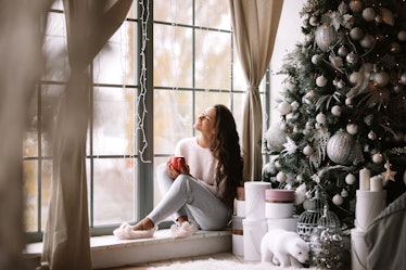 Charming dark-haired girl dressed in pants, sweater and warm slippers holds a red cup sitting on the...