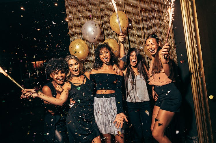 A group of girls laugh and pose in front of a gold backdrop at a club while confetti falls on New Ye...