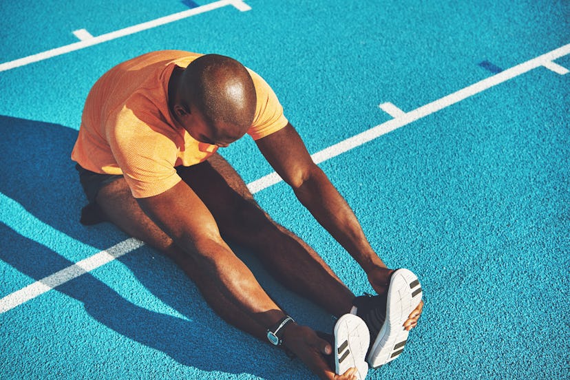 Focused young African male athlete in sportswear sitting alone on the lanes of a race track stretchi...