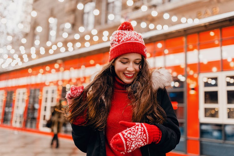 young pretty smiling happy woman in red mittens and knitted hat wearing winter coat walking in city ...