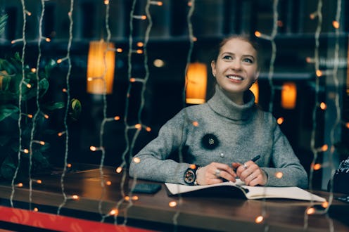 Young charming female smiling and looking outside window, writing Christmas wish-list and thinking o...