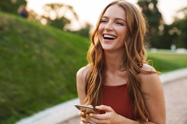 Photo of a happy laughing emotional young ginger woman walking in nature green park outdoors using m...