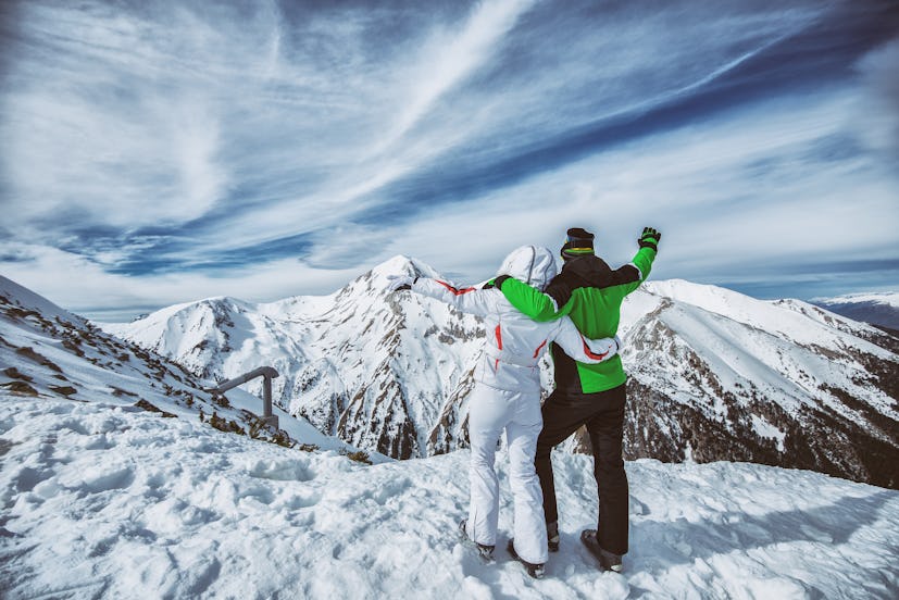 Couple in ski suits standing on a mountain peak, enjoying the view.