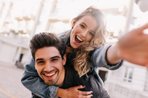 Curly adorable girl fooling around with boyfriend. Laughing graceful lady making selfie on street ba...