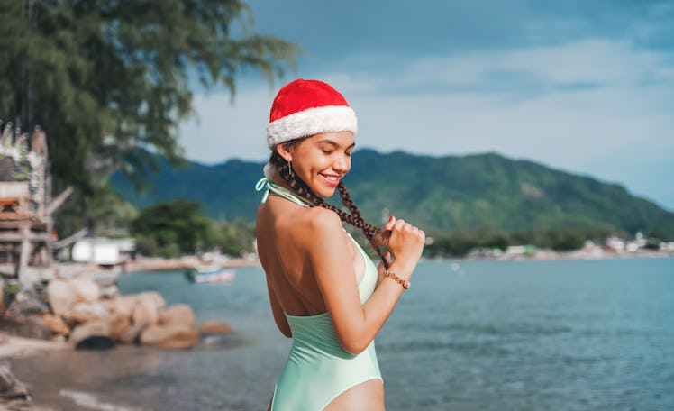 A woman with braids in a Santa hat and green bathing suit poses by the ocean on a Christmas vacation...