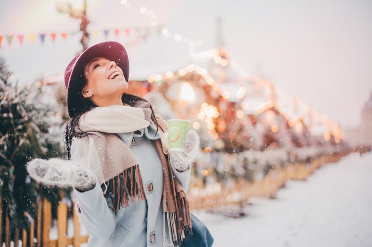 A happy woman stands in a snowy Connecticut Christmas town while holding a coffee mug and looking up...