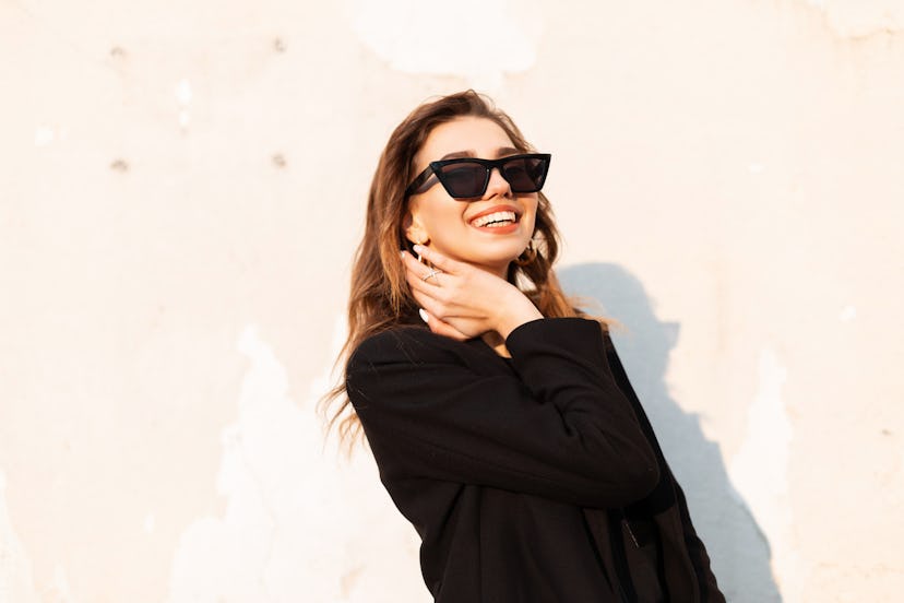 Pretty positive young hipster woman in black elegant coat in dark trendy sunglasses stands and smile...