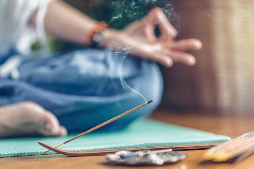Woman meditating in lotus position on turquoise yoga mat on wooden floor. Focus on incense stick and...