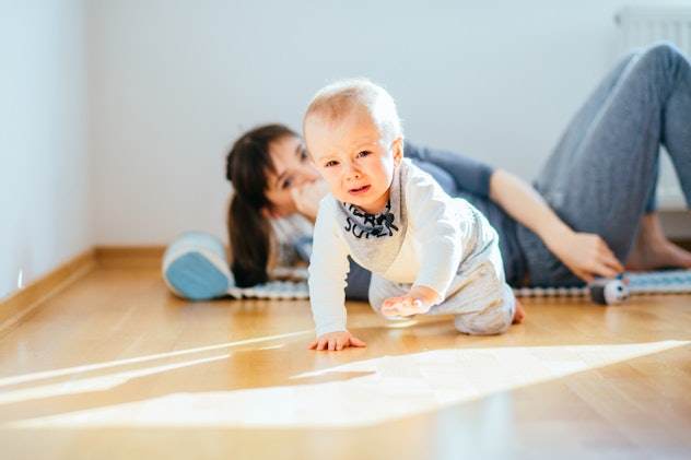 Crying infant baby boy crawling to the camera while his mother workout on mat on background behind h...