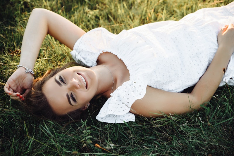 Happy smiling girl in a white summer sundress lies on the grass and looks into the camera. View from...