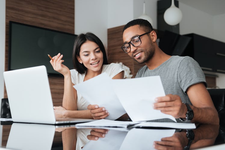Photo of cheerful loving young couple using laptop and analyzing their finances with documents. Look...