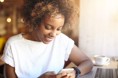 Adorable hipster dark-skinned woman with Afro hairstyle checking her news feed or messaging via soci...