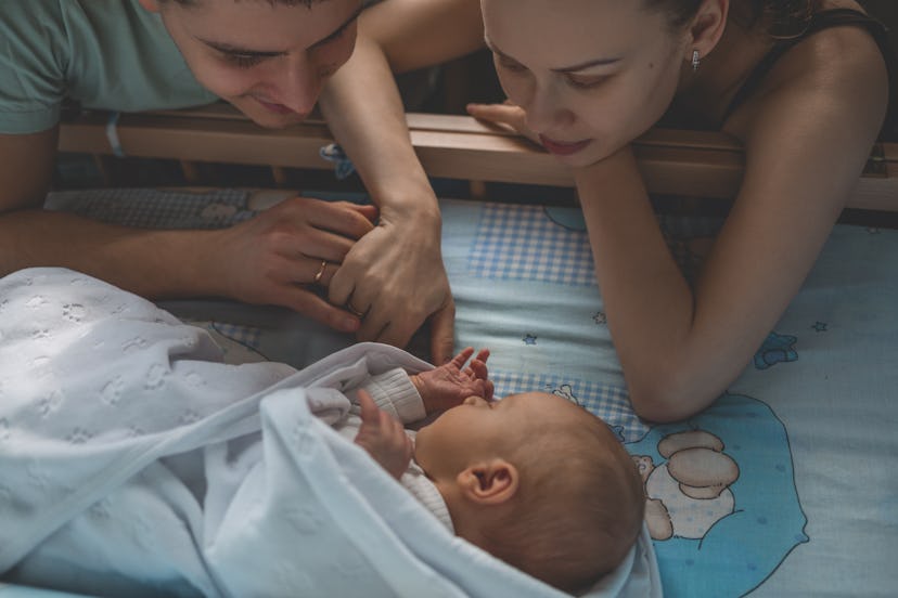 Woman and man look at newborn. Boy sleeps in his crib. Mom, dad and baby. Portrait of young family. ...