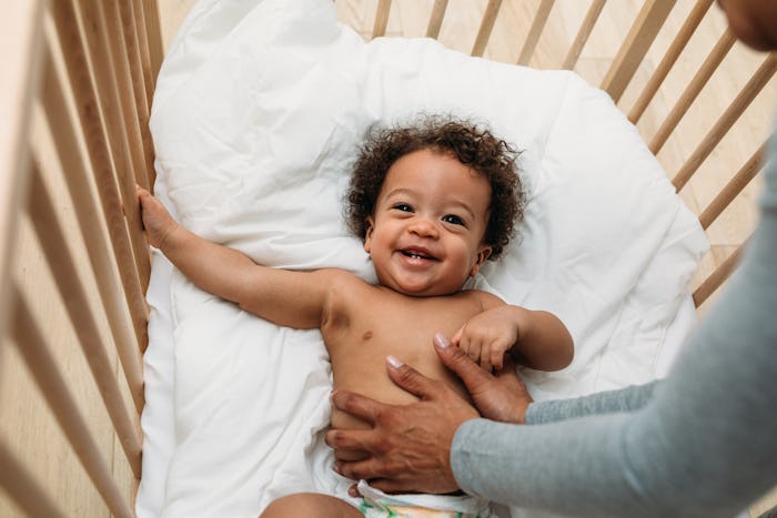 Happy baby boy in a crib. Cropped hands of mother tickling son lying on bed at home.