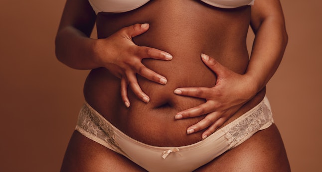 Captura recortada de mujer negra en ropa interior sosteniendo su vientre gordo.  Talla grande mujer en lencería sosteniendo su vientre con ambas manos sobre fondo marrón.