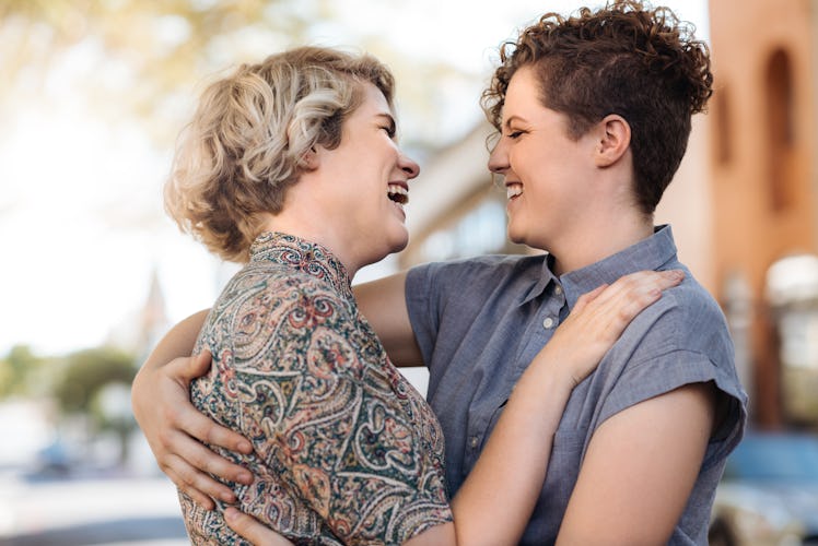 Laughing young lesbian couple standing arm in arm together on a city street listening to music on ea...