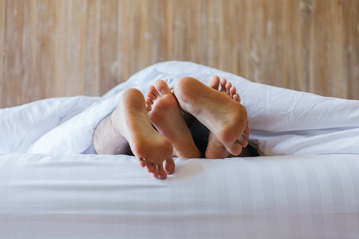 Close up of male and female feet on a white bed. The concept of loving couple having sex under white...