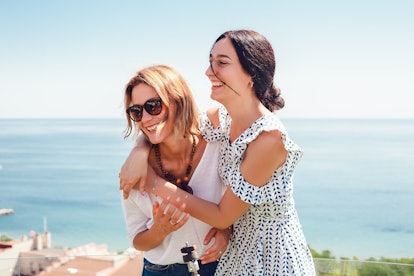 Two happy women hugging, standing on blue sky and sea background, stylish woman, bracelets, necklace...