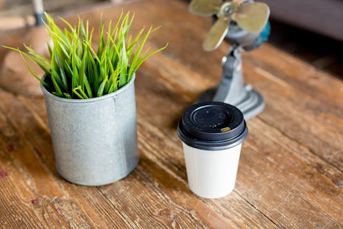 paper Cup of coffee on a wooden table next to a pot of green grass and light grey background on the ...