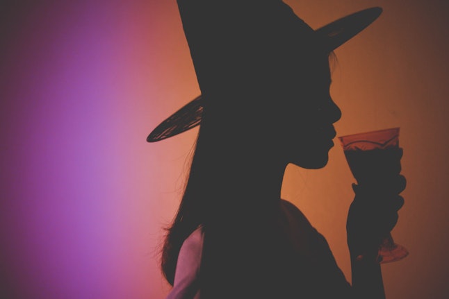Shadow image of a woman wearing a witch’s hat and holding a glass of drink with orange and purple light in the background. Halloween.