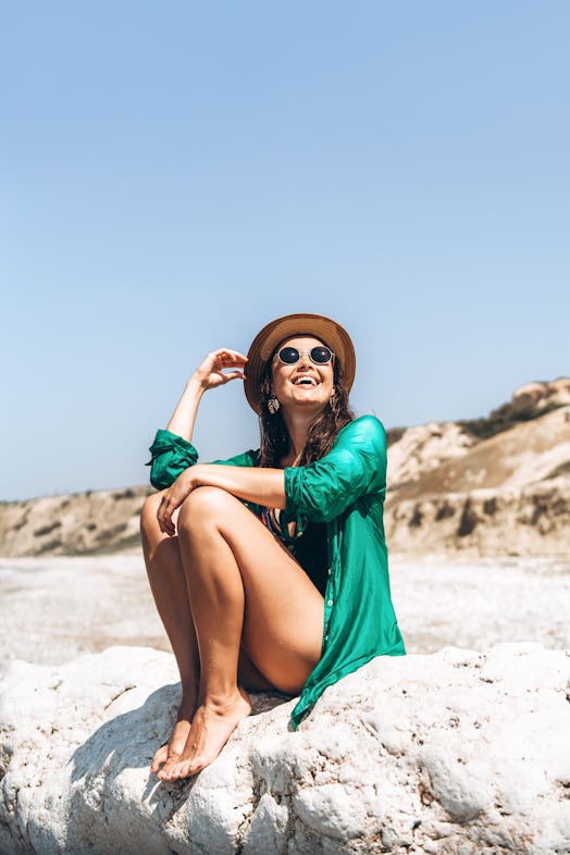 A stylish woman poses on a rock on a sandy beach while on vacation.