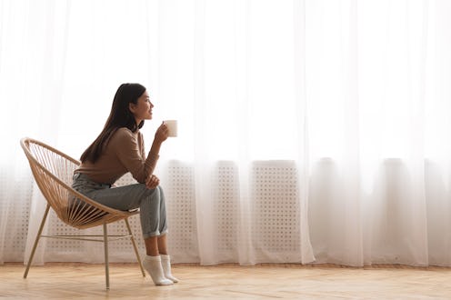 Young Girl Sitting In Modern Chair, Enjoying Coffee In Front Of Window, Side View