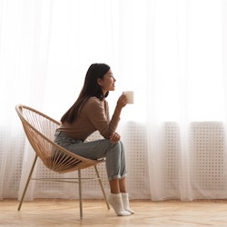 Young Girl Sitting In Modern Chair, Enjoying Coffee In Front Of Window, Side View