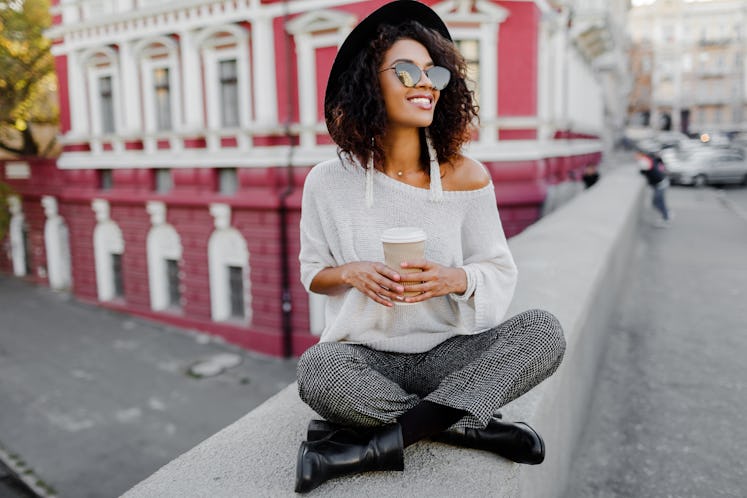 Street fashion look. Stylish black girl sitting on the bridge and holding cup of coffee or tea durin...
