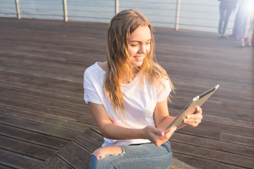 Happy smiling woman online ordering via portable touch pad while relaxing on embankment during recre...
