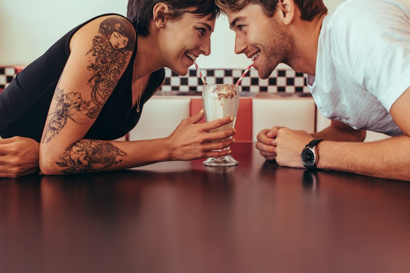 Close up of happy couple sharing a milk shake with straws sitting opposite to each other at a restau...