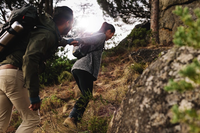 Female hiker helping her boyfriend uphill in the countryside. Young couple hiking in mountain.