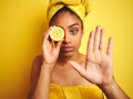 Afro woman wearing towel after shower holding slice lemon over isolated yellow background with open ...