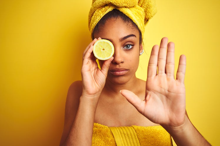 Afro woman wearing towel after shower holding slice lemon over isolated yellow background with open ...