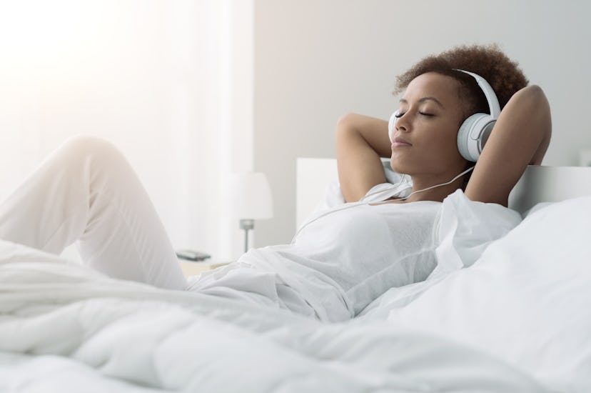 Young beautiful african american woman relaxing and listening to music using headphones, she is lyin...