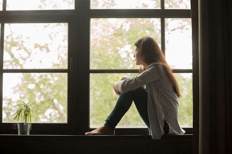 Thoughtful girl sitting on sill embracing knees looking at window, sad depressed teenager spending t...